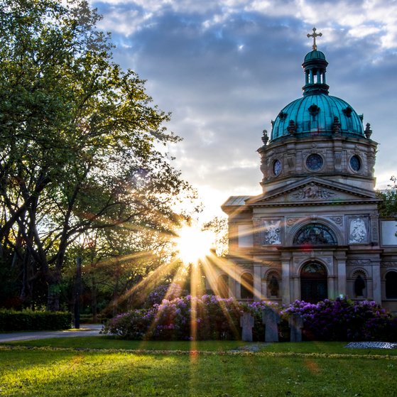 Sonnenuntergang im Freien in einem Park, eine Kapelle ist zu sehen, hinter der die Sonne in Strahlen untergeht