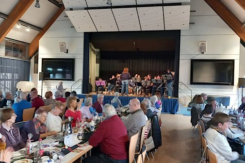 Panorama des Festsaals mit mehreren Tischreihen, an denen viele Leute sitzen. Im Hintergrund die Bühne mit einem Orchester.