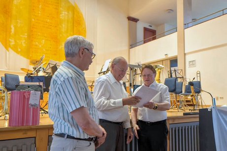 3 gut gekleidete, ältere Herren. Der Mann ganz rechts überreicht dem mittleren Mann eine Urkunde.