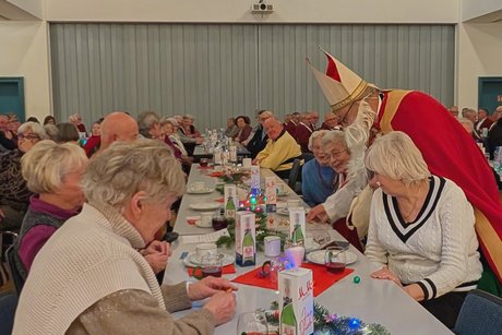 Vorweihnachtlicher Nachmittag Nikolaus beugt sich zwischen zwei Damen an einer Tischreihe und verteilt Süßigkeiten, während sich die Damen freuen.