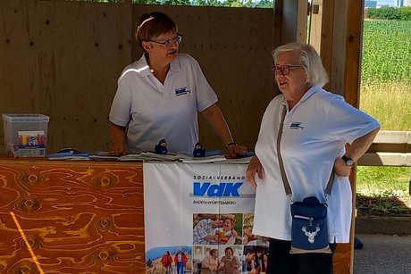 Zwei Frauen mit VdK-Poloshirts an einem Holzstand, eine steht davor, die andere dahinter. Am Stand hängt ein VdK-Stoffbanner.