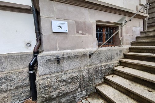 Wo geht's lang? Hinweistafel an der Rathausaußentreppe am Vordereingang auf den barrierefreien Hintereingang. Zu sehen ist auch eine nach unten verlaufende Regenrinne an der Außenwand sowie ein Kellerfenster