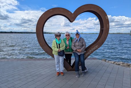 Drei &auml;ltere Damen stehen vor einer herzf&ouml;rmigen Metallskulptur am Ufer des Altm&uuml;hlsees, im Hintergrund der See.