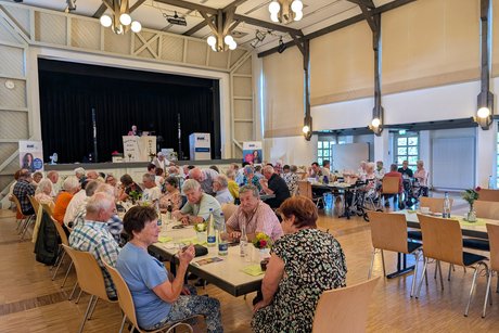 Panorama vom Festsaal mit mehreren Tischreihen, an denen ältere Menschen sitzen und sich unterhalten.