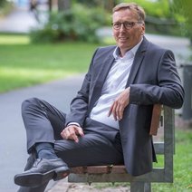 Ein sportlicher Herr mittleren Alters mit Brille sitzt l&auml;chelnd charmant im l&auml;ssigen, blauen Anzug auf einem auf einem Stuhl in der Natur