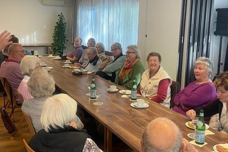 Ein langer Holztisch in einem hell beleuchteten Raum, um den mehrere Menschen &ndash; darunter &auml;ltere Erwachsene &ndash; sitzen und miteinander sprechen oder Getr&auml;nke trinken. Einige halten Kaffee-Tassen oder Wasserflaschen; auf dem Tisch sind auch Kuchenteller und ein St&uuml;ck Kuchen. Im Vordergrund ist der R&uuml;cken eines Mannes mit grauem Haar zu sehen. An den W&auml;nden h&auml;ngen Vorh&auml;nge vor einem Fenster, im Hintergrund ist eine Pflanze und eine T&uuml;r mit gr&uuml;nem Notausgangsschild sichtbar. Die Stimmung wirkt freundlich und gesellig, wie bei einem gemeinsamen Kaffeetrinken oder einer kleinen Veranstaltung.