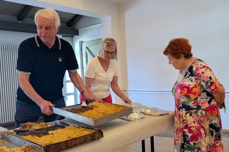 Tisch mit mehreren Blechkuchen. Hinter dem Tisch ein Mann und eine Frau, die die Kuchen verteilen. Davor eine Frau, die die Kuchen interessiert anschaut.