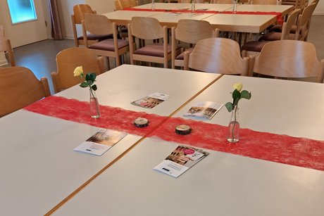 ansprechende, liebevolle Tischdeko Tisch mit rotem Tischband, einzelner Rose in einer kleinen Flasche, 2 kleinen Holzscheiben und Flyern
