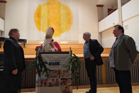 Vorweihnachtlicher Nachmittag Nikolaus am Rednerpult, drei Männer im Anzug blicken ihn an.