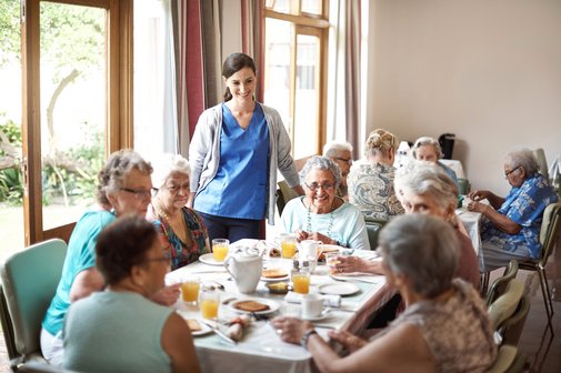 Esstisch in einem Seniorenheim, viele &auml;ltere Menschen sitzen zusammen am Tisch, unterhalten sich und essen. Am Tisch steht eine Pflegerin und erz&auml;hlt etwas oder h&ouml;rt gerade zu.