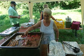 Die zweite Vorsitzende Agnes W. grillt Steaks und Würstchen unter einem Pavillion im Garten des Winzermuseums. Grillpfanne ist gut gefüllt. Ein Gast wartet auf seine Bestellung.