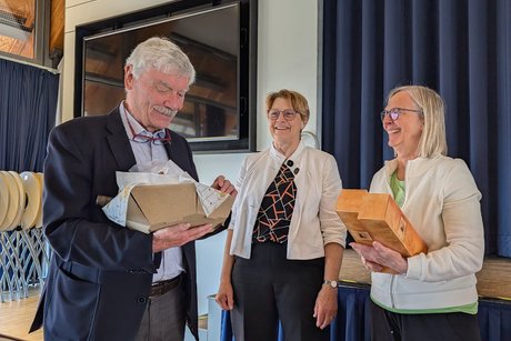 Ein Mann im Anzug schaut l&auml;chelnd in einen Geschenkkorb. Daneben stehen zwei Damen in schicken Blazern; eine von ihnen h&auml;lt zwei Geschenkboxen mit Weinflaschen. Alle Personen l&auml;cheln.