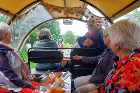 Blick von der Kutsche auf den Kutschbock; der Kutscher hat sich nach hinten gedreht und erzählt den Personen eine Anekdote. Die Personen auf den Sitzbänken links und rechts schauen ihm aufmerksam an.