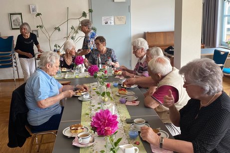 Sieben ältere Männer und Frauen sitzen gemeinsam an einem Tisch, essen Kuchen und unterhalten sich angeregt.