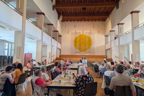 Panoramaaufnahme des Festsaals mit mehreren Tischreihen, auf denen Menschen sitzen und aufmerksam einem Mann im Hintergrund bei seiner Rede zuhören.