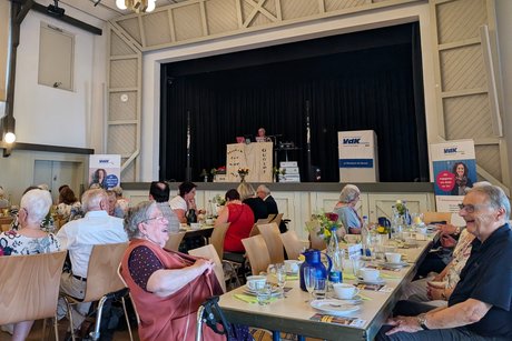Panorama vom Festsaal. Zentral eine Tischreihe, an der mehrere ältere Menschen sitzen. Im Vordergrund fällt besonders ein Mann und eine Frau auf, die sich gegenübersitzen und herzhaft lachen.