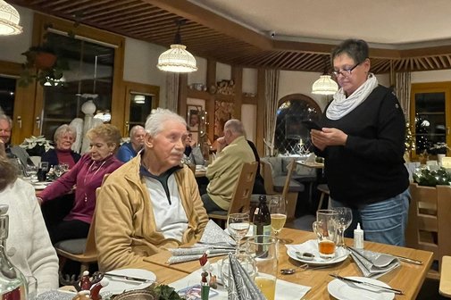 Beisitzerin Roswitha Philipp stehend und die Geschichte von "Omas Schurz" vortragend, an gedeckten Tischen sitzen die Gäste, ein Adventskranz mit einer brennenden Kerze im Hintergrund