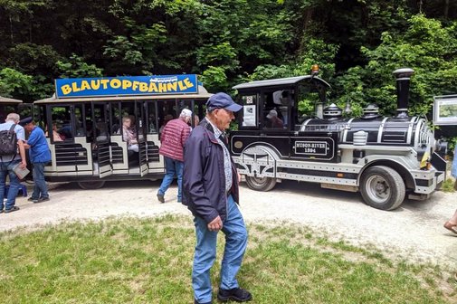 Mehrere Rentner stehen vor einer Touristenbahn, die wie eine kleine Dampflok aussieht; in den offenen Waggons des Zuges sitzen bereits mehrere Personen.