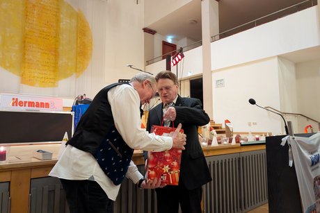 Ein Herr in schickem Anzug überreicht einem Mann in Weste ein Präsent.