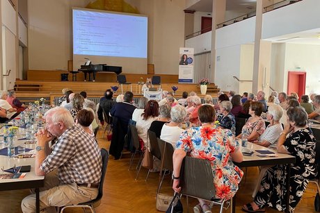 Panorama des Festsaals mit zwei Tischreihen, an denen ein bunt gemischtes Publikum sitzt und aufmerksam einem Vortrag lauscht.