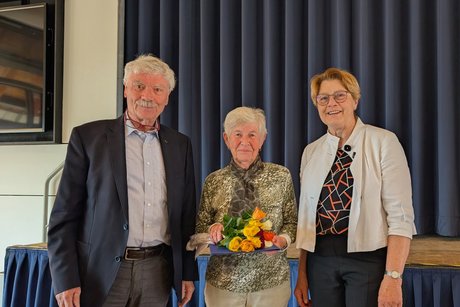 Gruppenfoto: Links ein Mann mit Schnauzbart, schick in Hemd und Anzug gekleidet, daneben eine Frau, die ein Blumengesteck h&auml;lt, und rechts eine Dame in einem modischen Blazer.