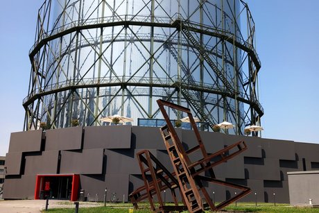 Metallfarbener, runder Turm mit modernem Flachbau ringsum und einem markanten, rot umrandeten Eingangsbereich.