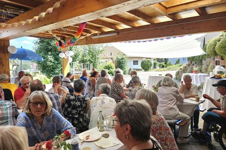 Viele Besucher sitzen an Tischen unter dem massiven Holzdach bei sch&ouml;nstem Wetter.
