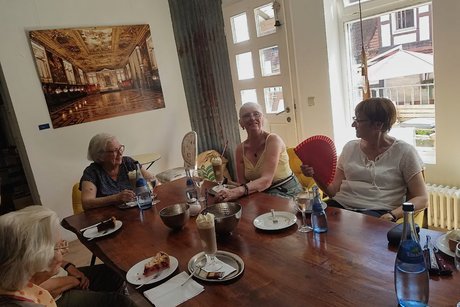 Fünf Frauen genießen gemeinsam Kaffee und Kuchen; hinter ihnen hängt ein großes Leinwandfoto eines goldenen Festsaals.