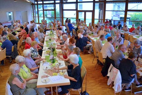 Panorama des Festsaals mit mehreren Tischen, an denen Menschen sitzen und vespern.