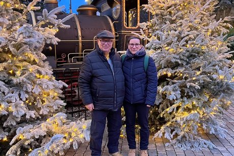 Zwei Mitglieder zwischen wei&szlig;en Tannenb&auml;umen und vor einem dekorativen Zug.
