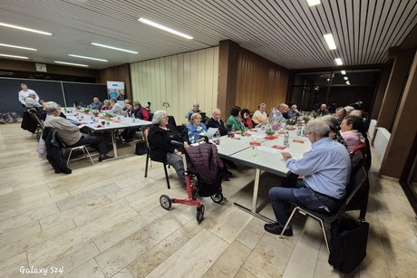 Mitglieder an L Förmig aufgestellten Tischen welche einem Redner zuhören.