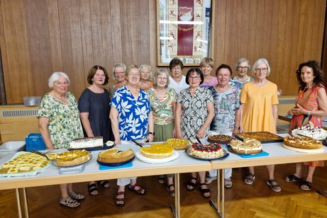 Vordergrund Tische mit 12 verschiedenen selbstgebackenen Kuchen, dahinter die Kuchenspenderinnen