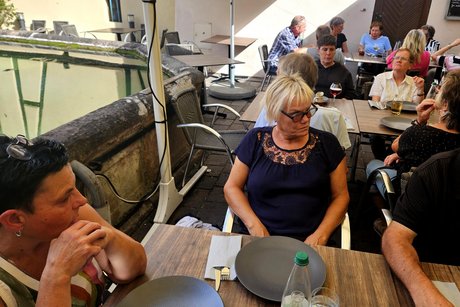 Alle sitzen im Freien an den gedeckten Tischen und warten auf die Flammkuchen. &Uuml;ber den B&auml;nken h&auml;ngen gro&szlig;e Sonnensegel.