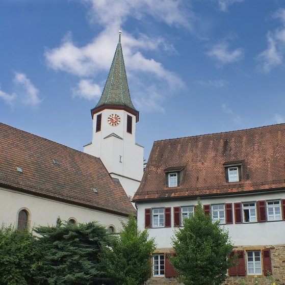 VdK OV Hochdorf Links das Kirchenschiff und der Kirchturm, daneben ein historisches Haus mit Natursteinmauer im Erdgeschoss und weiß verputztem Obergeschoss, vor blauem Himmel.