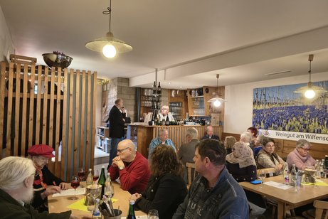 Gastraum mit zahlreichen sitzenden Personen an Tischen und der Wirtin an der Theke