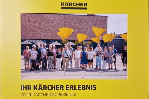 die 23 Teilnehmer der Werksführung bei der Firma Kärcher auf einem Gruppenfoto, das vor Beginn der Führung gemacht wurde. Die Personen stehen vor einem Werksgebäude in der Sonne. Im Hintergrund sind Sonnenschirme zu sehen.