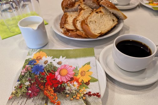 Blumenserviette auf einem Teller, daneben eine Tasse Kaffee, ein Milchkännchen und ein Teller mit frischem Hefezopf.
