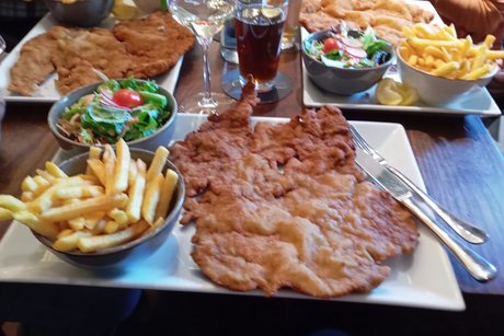 Ein gut gedeckter Mittagstisch mit riesigen Schnitzel, Schalen mit Pommes Frites, Salat und Getr&auml;nken.