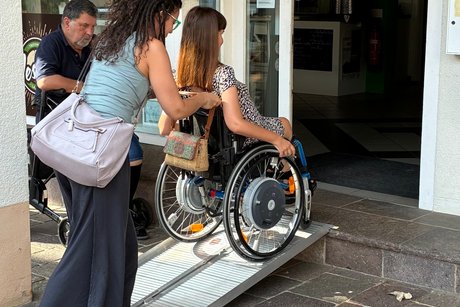 Unter Beobachtung von Vorsitzenden J&uuml;rgen Heiner nutzt Nora Welsch gemeinsam mit ihrer pers&ouml;nlichen Assistentin die mobile Rampe am Eingang des Caf&eacute; Wesley&acute;s.