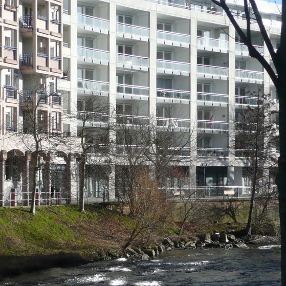 Unsere ansprechenden Räumlichkeiten finden Sie in der Pforzheimer Innenstadt neben der Seniorenresidenz Ambiente direkt am Enzufer Im Vordergrund sieht man die Enz mit ein paar Bäumen, im Hintergrund das Gebäude der Seniorenresidenz Ambiente und daneben das Gebäude, in dem unsere VdK Geschäftsstelle ist
