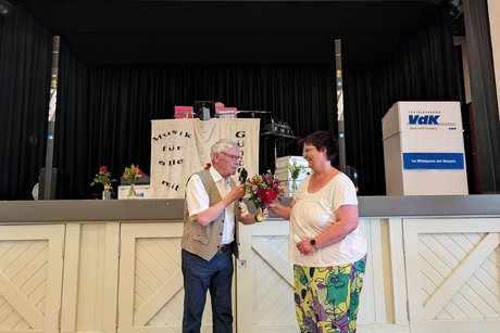 Ein älterer Herr im weißen Kurzarmhemd und Weste überreicht einer Frau einen bunten Blumenstrauß.