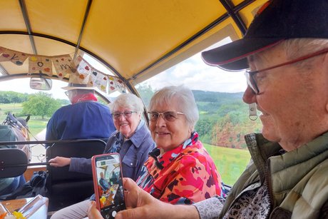 Im Vordergrund ein älterer Mann, der ein Foto von den beiden neben ihm sitzenden Damen macht.