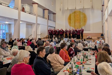 Vorweihnachtlicher Nachmittag Festsaal mit mehreren Tischreihen, an denen Menschen dicht nebeneinander sitzen, im Hintergrund Bühne mit Chor.