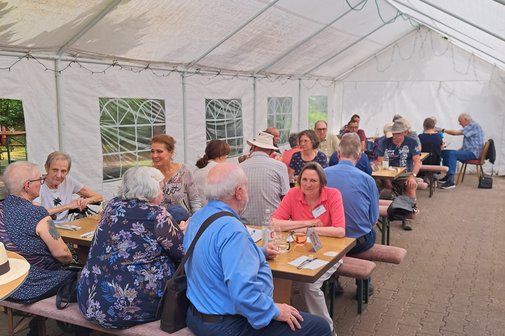 Ungef&auml;hr 30 Leute sitzen in einem halboffenen Zelt im Besen beim Essen jnd Sonnenschein umd unterhalten sich angeregt