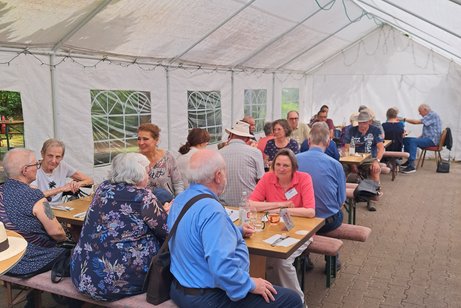 Ungef&auml;hr 30 Leute sitzen in einem halboffenen Zelt im Besen beim Essen jnd Sonnenschein umd unterhalten sich angeregt