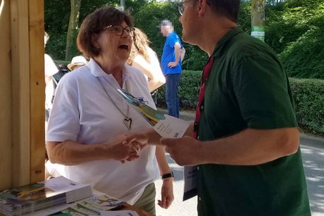 Vor dem VdK-Stand links steht eine Frau mit VdK-Poloshirt, die herzlich lacht, ihr gegenüber steht ein Mann im grünen Shirt.
