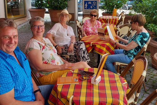 Fünf Personen an einem Tisch mit bunter Tischdecke; vier lächeln in die Kamera, eine Person schaut konzentriert in die Eiskarte.