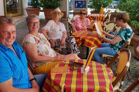 Fünf Personen an einem Tisch mit bunter Tischdecke; vier lächeln in die Kamera, eine Person schaut konzentriert in die Eiskarte.