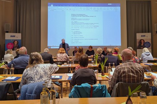 Blick in den Vortragssaal von hinten auf mehrere Tischreihen mit Konferenzteilnehmenden; vorne zeigt eine Leinwand die Tagesordnungspunkte, davor sitzt der geschäftsführende Vorstand des Kreisverbands.