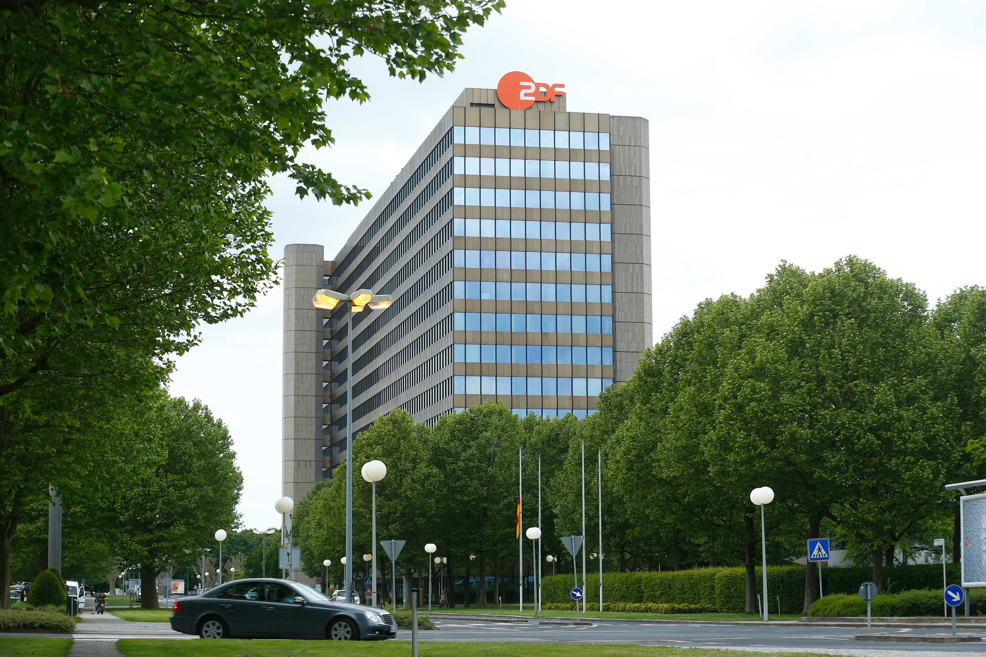 Hochhausgeb&auml;ude des ZDF mit Logo oben, im Vordergrund B&auml;ume und Stra&szlig;e mit 1 Pkw